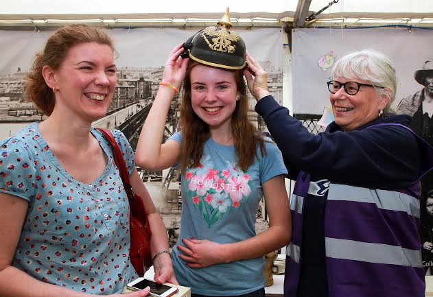 Events volunteer Karen Howe, slips a WWI German helmet onto the head of Edel Keogh, watched by her mum Shelagh, on their visit to the WWI memorabilia stand in the Festival Village tent.